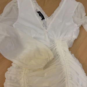 Vit kortklänning  - Supersöt vit kortklänning från Parisian med långa ärmar, v-ringning och rynkade detaljer längs hela klänningen. Klänningen har dragkedja i sidan och volangkant vid halsen. Materialet är lätt och känns luftigt, perfekt för sommarens alla tillfällen. Använts bara 2 gånger och ser ny ut. Köpt från boozt för 450kr. Pris kan diskuteras.