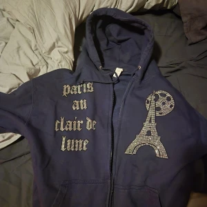 Mörkblå hoodie med strass från Blinglucid - Säljer en mörkblå hoodie från Blinglucid med strassdetaljer. Framsidan har texten 'Paris au clair de lune' i glittriga stenar och en stor Eiffel-torn-design med måne. Tröjan har huva, snörning och dragkedja. Perfekt för dig som gillar bling och Paris vibes.
