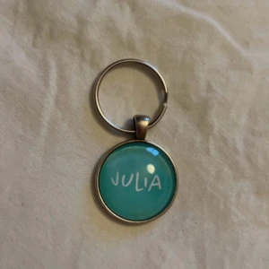 Nyckelring med turkos bricka Julia - Säljer en rund nyckelring med turkos bricka där det står 'JULIA' i vit text. Baksidan har ett snyggt geometriskt mönster i silverfärgad metall. Perfekt accessoar för dig som vill ha något personligt och färgglatt på nyckelknippan.