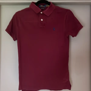 Vinröd Polo Ralph Lauren t-shirt XS - Snygg vinröd t-shirt från Polo Ralph Lauren i slim fit-modell. Klassisk pikékrage med två vita knappar och blå broderad logga på bröstet. Tillverkad i mjuk bomull som känns skön mot huden. Perfekt för dig som gillar stilrena och tidlösa plagg.
