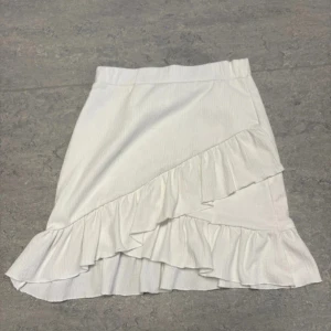 Vit volangkjol från NLY Trend - Supersöt vit kortkjol från NLY Trend med volangdetaljer och omlottkänsla framtill. Kjolen är gjord i mjuk polyester med lite stretch för skön passform. Perfekt att styla med sneakers eller sandaler för en fräsch look.