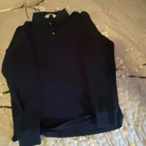 Mörkblå pikétröja med lång ärm - Snygg mörkblå pikétröja med klassisk krage och två knappar framtill. Tröjan har lång ärm och är tillverkad i ett mjukt bomullsmaterial som känns skönt mot huden. Perfekt för dig som gillar stilrena och enkla plagg med lite sportig vibe.