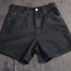 Svarta högmidjade jeansshorts - Svarta jeansshorts från Gina Tricot med hög midja och klassisk femficksdesign. Modellen har raka ben och knappstängning framtill. Materialet är robust denim som ger en cool och avslappnad vibe. Perfekt för dig som gillar enkel och stilren look.