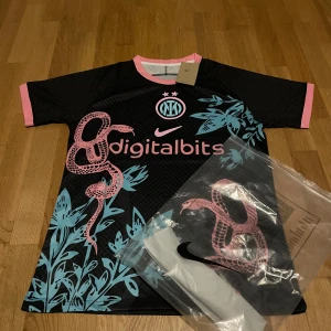 Inter Milan Dri-Fit fotbollströja Nike - Inter Milan Dri-Fit fotbollströja från Nike med svart bas, rosa detaljer och blått tryck. Coolt ormmotiv och bladmönster på framsidan, samt Digitalbits-logga. Kortärmad med rosa krage och ärmslut. Tillverkad i lätt, ventilerande polyester.