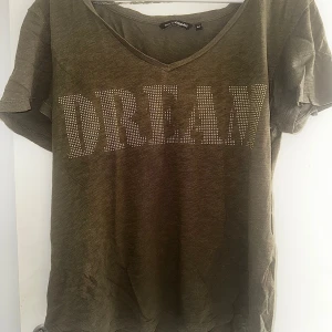 Olivgrön t-shirt DREAM - Olivgrön t-shirt från KappAhl i 100% bomull med v-ringning och korta ärmar. Framsidan har ett stort DREAM-tryck i glittriga nitar. Passformen är normal och tyget är mjukt och skönt mot huden.