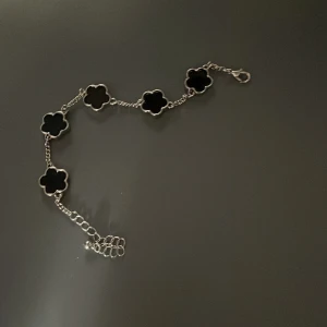 Silverarmband med svarta blommor - Stilrent armband i silverfärg med fem svarta blommor i klöverform längs kedjan. Perfekt accessoar för att lyfta din outfit med en cool och elegant touch. Justerbar längd med karbinlås. Ca 21 cm 