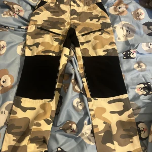 Camo täckbyxor från 157 Funk, strl 110 (barn) - Säljer ett par täckbyxor från 157 Funk i camomönster med beige, bruna och svarta toner. Byxorna har förstärkta svarta partier på knän och bak, dragkedjor vid fickorna och justerbar midja. Perfekta för vinteraktiviteter och lek utomhus.