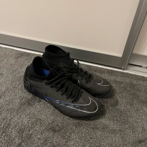 Nike Mercurial svarta fotbollsskor - Säljer ett par Nike Mercurial fotbollsskor i svart med blå och vita detaljer. Skorna har snörning, högre strumpliknande skaft och blå sula med fasta dobbar. Tillverkade i syntetmaterial för bästa grepp och känsla på planen. Skicket är som nytt! Endast använd i en halv säsong. Kommer utan orginalbox då den är slängd.