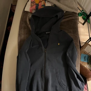 Mörkblå hoodie från Lyle & Scott - Säljer en mörkblå hoodie från Lyle & Scott med dragkedja och huva. Tröjan har två fickor framtill och den klassiska gula fågel-loggan på bröstet. Perfekt för chill dagar och enkel att matcha med jeans eller joggers.