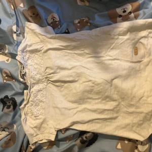 Vit t-shirt med spets från Hampton Republic (barnkläder) - Vit t-shirt från Hampton Republic med korta ärmar och snygga spetsdetaljer upptill. T-shirten har rund hals och ett litet metallmärke framtill. Materialet känns mjukt och skönt, perfekt för chill dagar. Spetsen har broderade blommor som ger en extra touch.