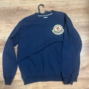 Mörkblå sweatshirt från Moncler - Snygg mörkblå sweatshirt från Moncler med stort broderat logomärke på bröstet. Tröjan har rund hals, långa ärmar och ribbade muddar vid ärmslut och nederkant. Perfekt för dig som gillar stilrena och exklusiva plagg.