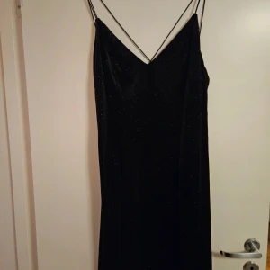 Svart glittrig klänning H&M - Supersnygg svart klänning från H&M med tunna axelband som korsas i ryggen. Klänningen har ett glittrigt tyg som ger en lyxig känsla och en v-ringad front. Perfekt för dig som vill sticka ut med stilren elegans och coola detaljer.