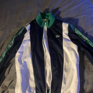 Lacoste sport kofta - Snygg kofta från Lacoste Sport med breda vita och mörkblå paneler, gröna detaljer vid krage och midja samt stora Lacoste-loggor längs ärmarna. Koftan har dragkedja framtill och klassisk krokodil-logga på bröstet. Perfekt för en sportig och clean look.