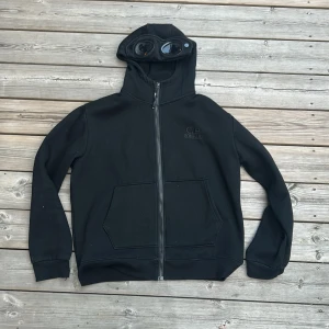 C.P. Company tröja - Svart hoodiejacka från C.P. Company med ikoniska goggles i huvan. Jackan har dragkedja framtill, två stora fickor och broderad logga på bröstet. Tillverkad i mjuk bomull, perfekt för en streetwear-look. Inga fel.