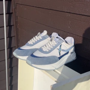 Blå och vita sneakers från Axel Arigato - Snygga sneakers från Axel Arigato i vitt och ljusblått med broderad fågel på sidan. Skorna har rund tå, platt sula och snörning. Ovandelen är i mocka och textil, vilket ger en fräsch och modern vibe. Perfekta för dig som gillar stilrena detaljer och cool design.