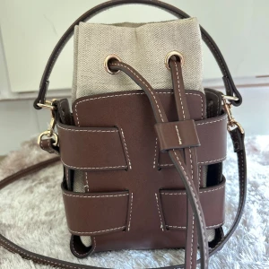 Brun bucket bag från H&M - Säljer den efterfrågade Snygga bucket bag från H&M i brun skinnimitation med kontrasterande vita sömmar och beige canvasinsats. Väskan har både handtag och axelrem samt cool snörning upptill. Perfekt för dig som vill ha en trendig och praktisk väska med unik design. 