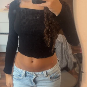 Svart croppad långärmad topp - Säljer en svart croppad topp med lång ärm och fyrkantig halsringning. Toppen har en smockad, ribbad struktur som ger en snygg och bekväm passform. Perfekt att matcha med jeans eller kjol för en cool och trendig look.