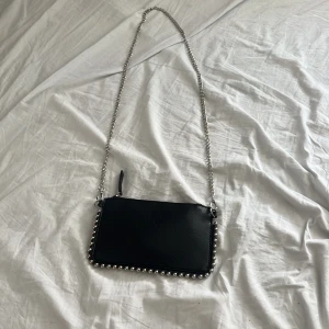 Svart axelväska med kedja och nitar - Snygg svart axelväska i skinnimitation med silverfärgad kedja som axelrem och coola nitar längs kanterna. Väskan har minimalistisk design och stängs med dragkedja upptill. Perfekt för dig som gillar edgy detaljer och stilren look.