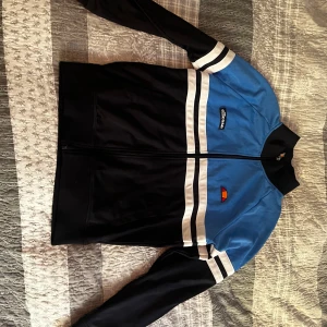 Ellesse blå och svart vindjacka M - Snygg ziptröja från Ellesse i blått och svart med vita ränder över bröstet och ärmarna. Tröjan har dragkedja hela vägen, ståkrage och Ellesse-logga på bröstet. Perfekt för en sportig och retro look.