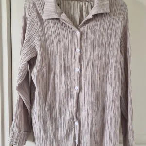 Beige crinkle-skjorta med byxor  - Snygg beige skjorta med krinklat tyg och klassisk krage. Skjortan har vita knappar framtill och långa ärmar med breda muddar. Materialet är mjukt och luftigt, perfekt för en chill och avslappnad stil. Passar dig som gillar enkel men cool design. Finns en knapp är fattas 