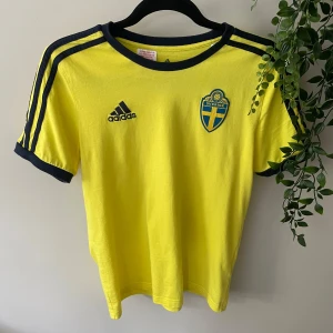 Sverige tröja 🇸🇪 - Gul fotbollströja från Adidas med blå detaljer och Sveriges landslagsemblem på bröstet. Klassiska Adidas-ränder på axlarna och blå kant vid hals och ärmslut. Materialet är mjukt och lätt, perfekt för match. Det står L i tröjan men det är i us storlek. 