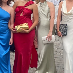 Röd långklänning med omlott och slits - Säljer en röd långklänning i glansigt material med snygg omlott-detalj och cutout framtill. Klänningen har en axelbandslös look med asymmetrisk axel och en slits på sidan för extra edge. Perfekt för dig som vill sticka ut med en cool och trendig stil.