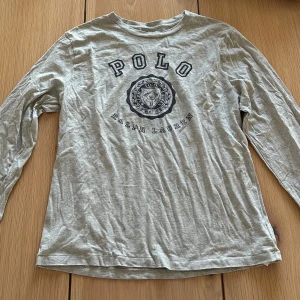 Grå långärmad tröja från Polo Ralph Lauren - Säljer en grå långärmad tröja från Polo Ralph Lauren med stort tryck på bröstet och text på ena ärmen. Tröjan är i mjuk bomull och har en avslappnad passform. Perfekt för chill dagar och snygg till jeans.