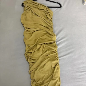 Guld one shoulder klänning PrettyLittleThing - Snygg guldglänsande kortklänning från PrettyLittleThing med one shoulder-design och draperade detaljer. Klänningen har en tight passform och är tillverkad i ett mjukt, glansigt syntetmaterial som ger en lyxig känsla. Perfekt för dig som vill sticka ut med en cool och trendig look. Oanvänd då den var för liten i storleken