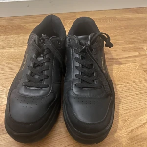 Svarta sneakers i skinn med snörning - Stilrena svarta sneakers med lågt skaft och klassisk design. Skorna har ovandel i skinn, svarta snören och diskret perforering på tån. På sidan finns en guldfärgad textdetalj med siffror. Perfekta för en clean och urban look.