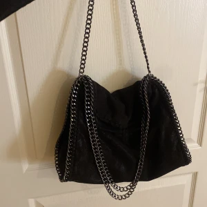 Svart axelväska med kedjedetaljer - Cool svart axelväska med tunga silverfärgade kedjor längs kanterna och som axelrem. Väskan har en mjuk, slouchy form och insidan är fodrad med leopardmönstrat tyg i bruna toner. Perfekt för dig som gillar edgy detaljer och djurmönster.