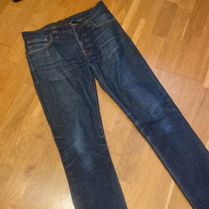 Mörkblå jeans från Nudie Jeans - Säljer ett par klassiska mörkblå grim tim jeans från Nudie Jeans med snygga kontrastsömmar och ikoniska bakfickor med vågigt mönster. Jeansen har rak passform och är tillverkade i slitstarkt bomullsdenim. Perfekta för dig som gillar stilrena och tidlösa jeans. Enda problemet är ett litet hål i fickan som inte syns när man har på sig dom! 30/31 står 32/34 men små i storleken!