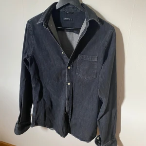 Grå jeansskjorta från J.Lindeberg - Snygg grå jeansskjorta från J.Lindeberg med klassisk krage, bröstficka och vita knappar. Skjortan har lång ärm och är tillverkad i ett robust jeanstyg som ger en cool och avslappnad look. Perfekt att bära öppen eller stängd över en t-shirt.