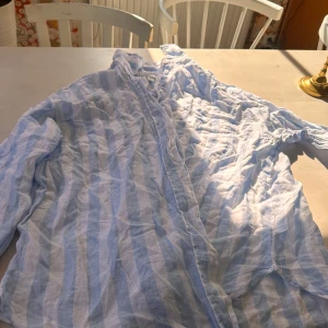 Randig linneskjorta från H&M, S - Säljer en luftig och snygg skjorta från H&M i storlek S. Skjortan är tillverkad i 100% linne och har ett fräscht randigt mönster i ljusblått och vitt. Perfekt för varma dagar och har långa ärmar samt knappar framtill.