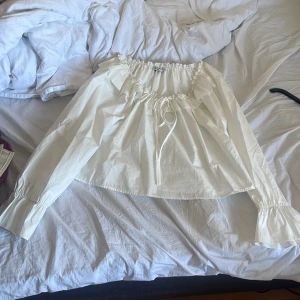 Vit offshoulder blus från NA-KD - Supersöt vit blus från NA-KD med offshoulder-modell och långa ärmar. Blusen har volangdetaljer runt halsen och knytning framtill. Tillverkad i bomull, perfekt för en fräsch och trendig look.