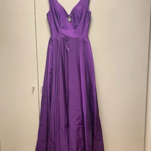 Lila långklänning med v-ringning - Säljer en lila långklänning i glansigt satinmaterial med v-ringad front och en stor kristall som detalj vid urringningen. Klänningen är ärmlös och har en dragkedja i ryggen. Perfekt för bal eller festliga tillfällen när du vill sticka ut. Priset kan diskuteras 