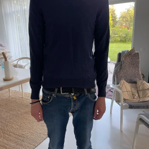 J.Lindeberg Mörkblå stickad tröja herr - Snygg mörkblå stickad tröja från J.Lindeberg med rund hals och långa ärmar. Tröjan har en klassisk passform och ribbade muddar vid ärmslut och nederkant med ett litet j.lindeberg märke av samma färg. Perfekt för dig som gillar stilrena och enkla plagg. Passar till jeans eller chinos. Modellen är 190cm och väger 69kg