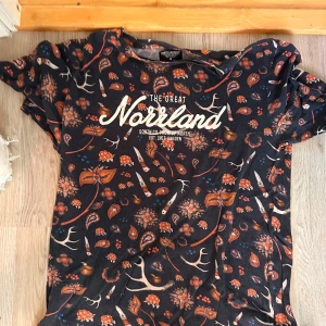 Mönstrad t-shirt från SQRTN Company - Snygg t-shirt från SQRTN Company med coolt tryck 'The Great Norrland' på bröstet. Svart botten med orange och beige blommor, knivar och horn i mönstret. Rund hals och korta ärmar, perfekt för dig som gillar streetwear med unik design.