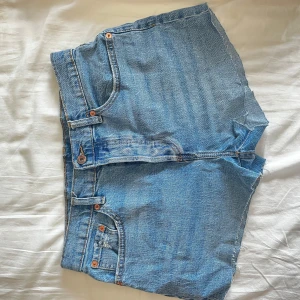Blå jeansshorts med rå kant - Säljer ett par klassiska blå jeansshorts från 157 med rå, avklippt kant och fem fickor. Shortsen har normal passform och midwaist, med snygga kopparfärgade nitar och knapp. Perfekta för sommaren och chill häng.