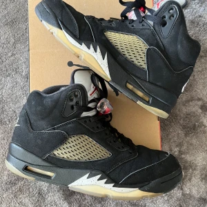 Nike Air Jordan 5 OG metallic  - Säljer ett par Nike Air Jordan 5 Retro i svart mocka med meshpaneler i beige och detaljer i vitt och rött. Skorna har högt skaft, snörning och ikoniska Nike Air-loggor på hälen. Sulan är genomskinlig med svarta detaljer och Jumpman-logga undertill.
