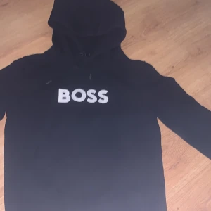 Svart hoodie från Hugo Boss med tryck - Svart hoodie från Hugo Boss i storlek S med stort vitt BOSS-tryck på bröstet. Tröjan har huva med snörning och långa ärmar. Materialet är mjuk bomull, perfekt för chill dagar. Klassisk och stilren design som funkar till jeans eller joggers. Pris kan diskuteras