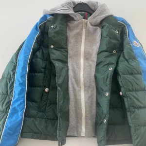 Moncler grön dunjacka  - Snygg grön dunjacka från Moncler med blå ränder längs ärmarna och vit piping. Jackan har en grå huva i sweatshirtmaterial och dragkedja framtill. Jackan är i väldigt bra skick. Jag skulle säga att jackan sitter ungefär 164-170Pris kan diskuteras vid snabb affär🫡