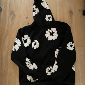 Svart hoodie med vita blommor Denim Tears - Svart hoodie från Denim Tears i storlek S med stora vita blommönster över hela tröjan. Klassisk huva och lång ärm, perfekt för dig som vill sticka ut med en unik och trendig look. Mjuk bomullskänsla och relaxed fit.