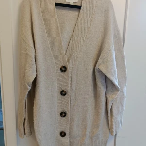 Beige oversized kofta med knappar - Säljer en beige oversized kofta med stora svarta knappar framtill. Koftan har långärmad design och bred ribbad kant vid halsen och längs knappslån. Perfekt för lager-på-lager och chill stil. Materialet känns mjukt och stickat.