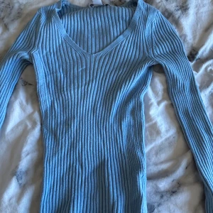 Ljusblå ribbad långärmad tröja - Säljer en ljusblå ribbad tröja med v-ringad hals och långa ärmar. Tröjan har en slim passform och är gjord i mjukt stickat material som sitter snyggt på kroppen. Perfekt för dig som gillar stilrena och enkla plagg med lite edge.