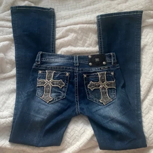 Miss me jeans - Snygga blå jeans från Miss Me med bootcut passform och coola broderade kors på bakfickorna. Jeansen har dekorativa nitar, tydliga kontrastsömmar och gnistrande knappar. Materialet är klassisk denim med lätt slitning för en trendig look.