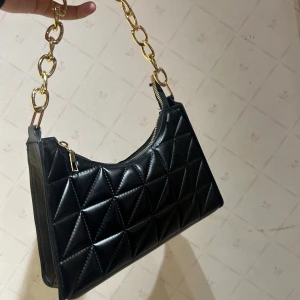 Svart quiltad axelväska med kedja - Snygg svart axelväska i quiltat skinnimitation med guldig kedja som axelrem. Väskan har geometriskt mönster med trianglar och dragkedja upptill. Perfekt accessoar för att lyfta din outfit med en lyxig touch.