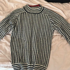 Randig stickad tröja från Pelle P - Säljer en randig stickad tröja från Pelle P i storlek L. Tröjan har svart och vit mönster med en hög krage som har en röd detalj upptill. Materialet är en mix av bomull, polyamid och merinoull. Långärmad och med en cool knappdetalj på axeln.