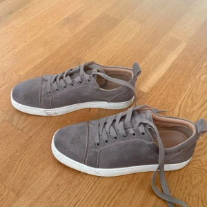 Grå louboutin skor - Snygga grå sneakers i mocka som liknar Louboutin med klassisk röd sula och vita kanter. Skorna har rund tå, platt sula och snörning. Insidan är ljusbeige och de har diskreta detaljer som ger en clean look. Perfekta för dig som gillar stilrena och exklusiva sneakers.