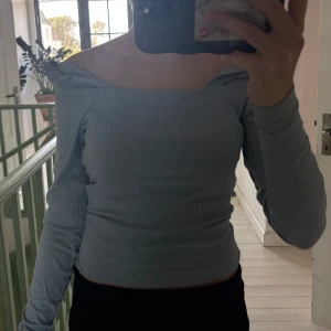 Ljusblå ribbad offshoulder topp - Supersnygg ljusblå ribbad topp med offshoulder och långa ärmar. Sitter tight och har en liten volangdetalj vid axlarna som ger en extra touch. Perfekt för dig som gillar en stilren men ändå trendig look.