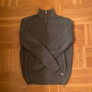 Grå ribbad half zip från ICON Amsterdam - Snygg grå ribbstickad tröja från ICON Amsterdam i storlek M. Tröjan har en hög krage med half zip-dragkedja och diskret metallogo vid nederkanten. Perfekt för lager-på-lager och chill stil under höst och vinter.